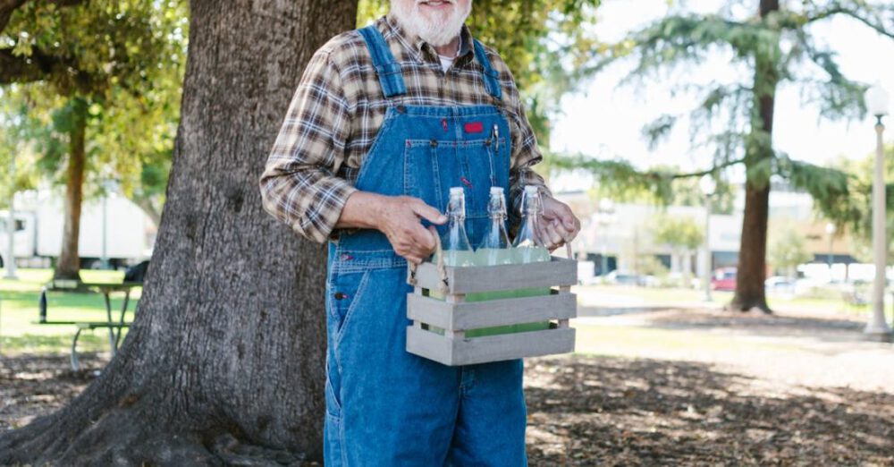 Cider - Farmer in Blue Denim Overalls and Fedora Hat Holding a Crate with Cider Bottles